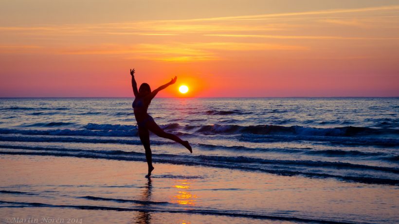 Een vrouw danst alleen op het strand, bij een ondergaande zon. Misschien is ze vrijgezel, misschien ook niet... Het ziet er hoe dan ook vrolijk uit.
