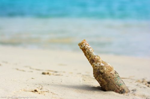 een oud bierflesje steekt uit het zand, op een mooi strand. Kristalhelder water op de achtergrond.